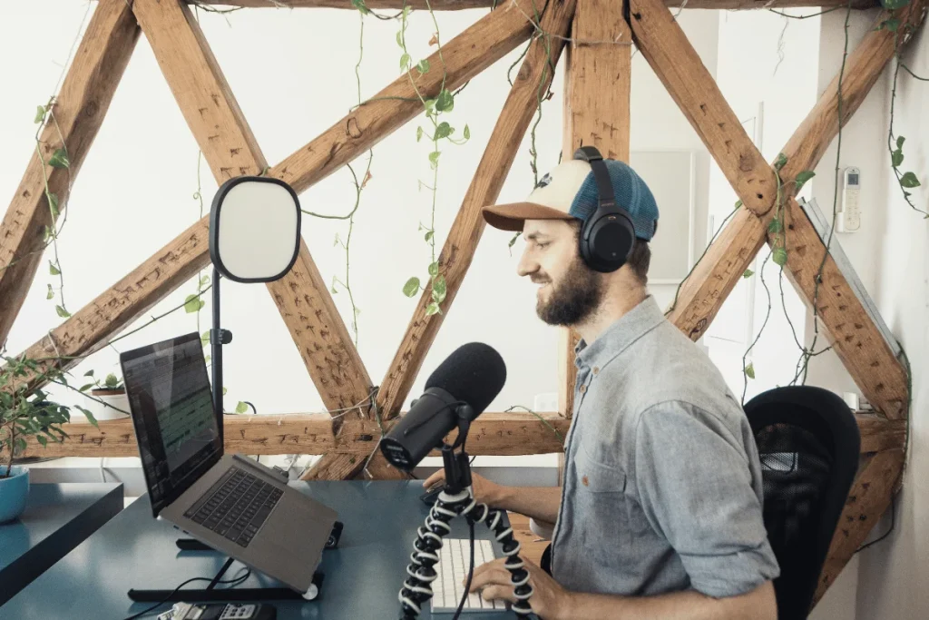 A bearded man in a denim shirt records a podcast in an airy studio with exposed wooden beams and lush plants.