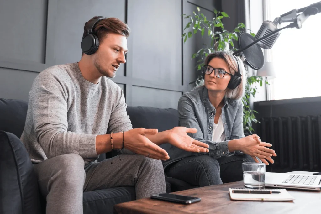 Two people with headphones gesturing during a podcast recording session.