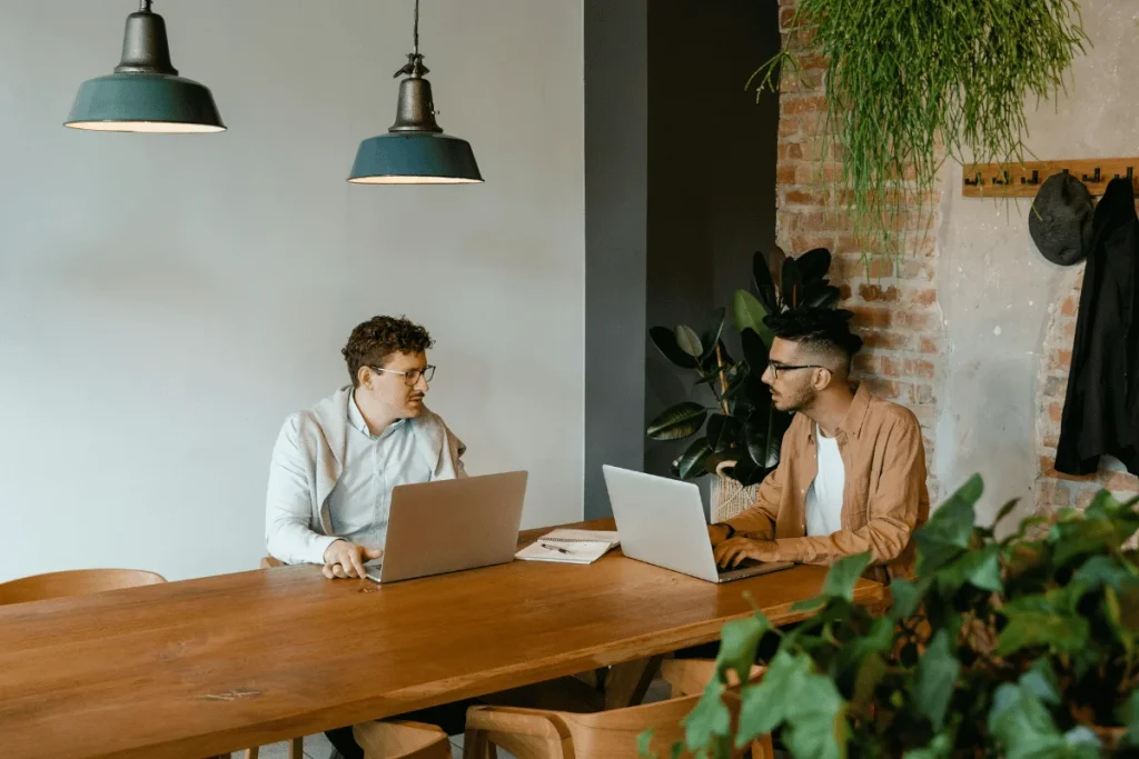 Two individuals sit at a wooden table in a modern office space, focused on their laptops with greenery in the background.