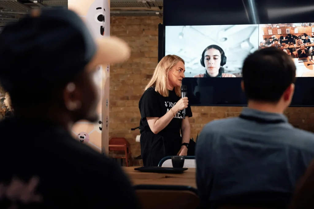 A speaker in a black shirt presenting with a microphone during a hybrid event with a video conference screen in the background.