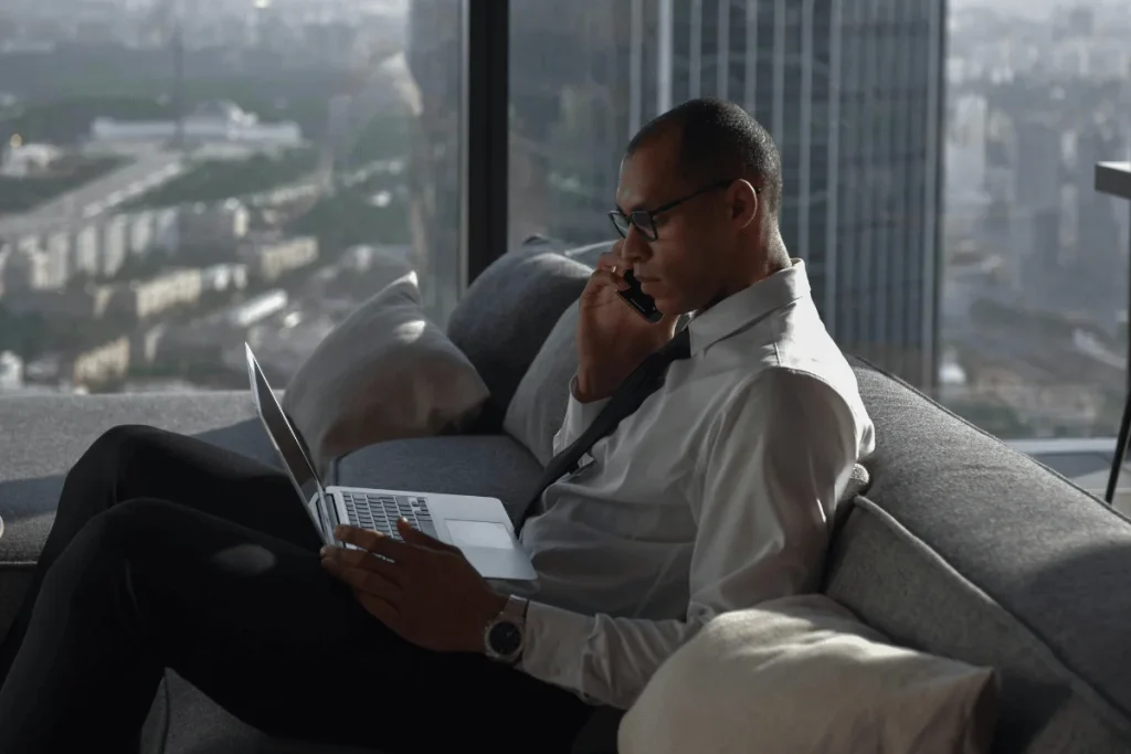 A professional man wearing a suit sits on a couch, talking on the phone while using a laptop, with a city view outside.