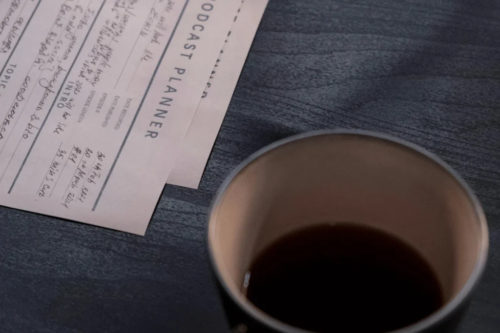 Detailed view of a podcast planner and a cup of coffee on a dark wooden table.