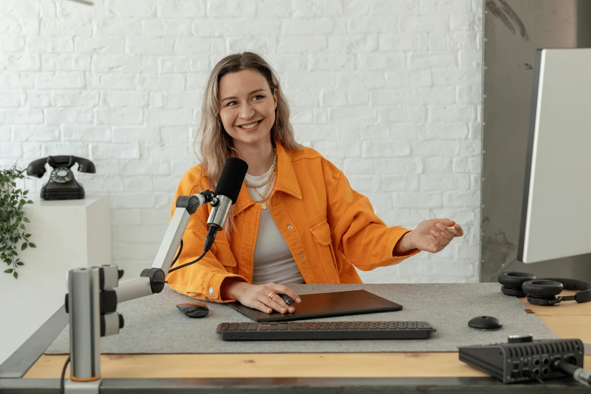 Person in an orange jacket sitting at a desk with a microphone and digital tablet.