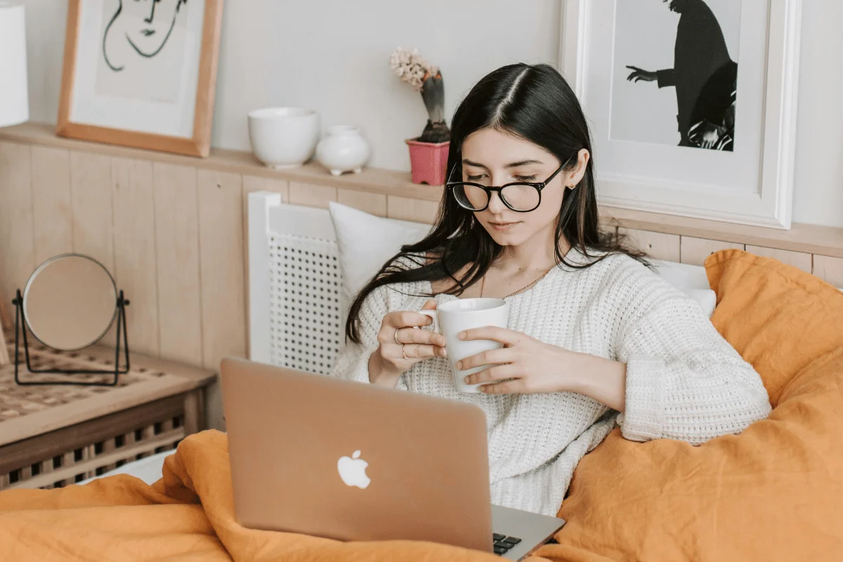 Young woman with glasses enjoying a cup of coffee while working on her laptop in bed.