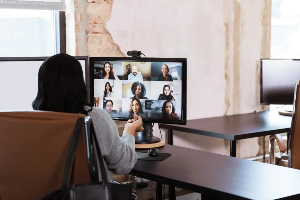 A professional attending a virtual meeting displayed on a monitor in a modern office setting with exposed brick walls.