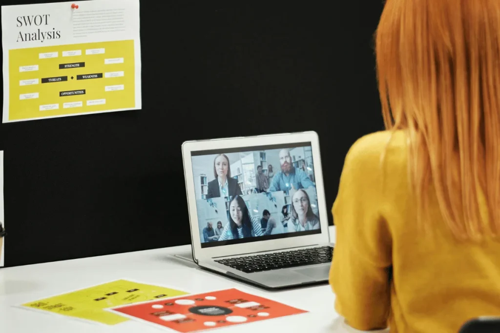 Person in a yellow top at a desk with a laptop showing a video call, SWOT Analysis chart on the wall.