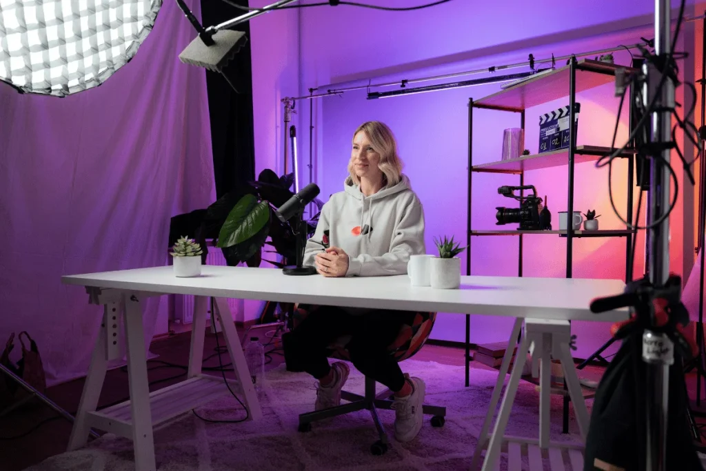 Person sitting at a desk in a video recording studio with lighting and camera equipment.