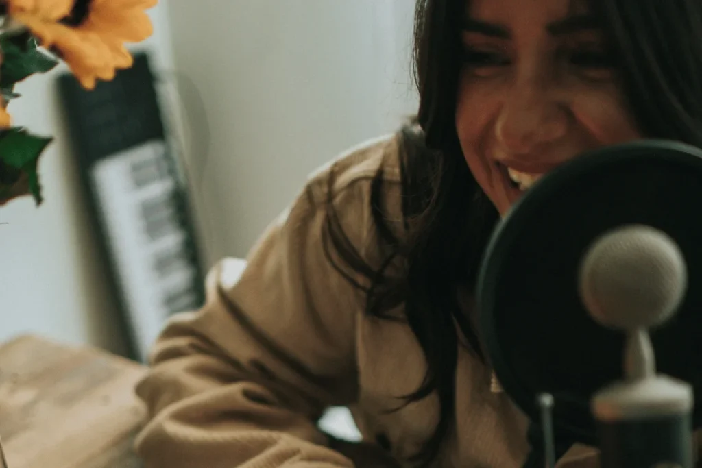 Person in a beige shirt with headphones in front of a microphone, possibly podcasting or recording.