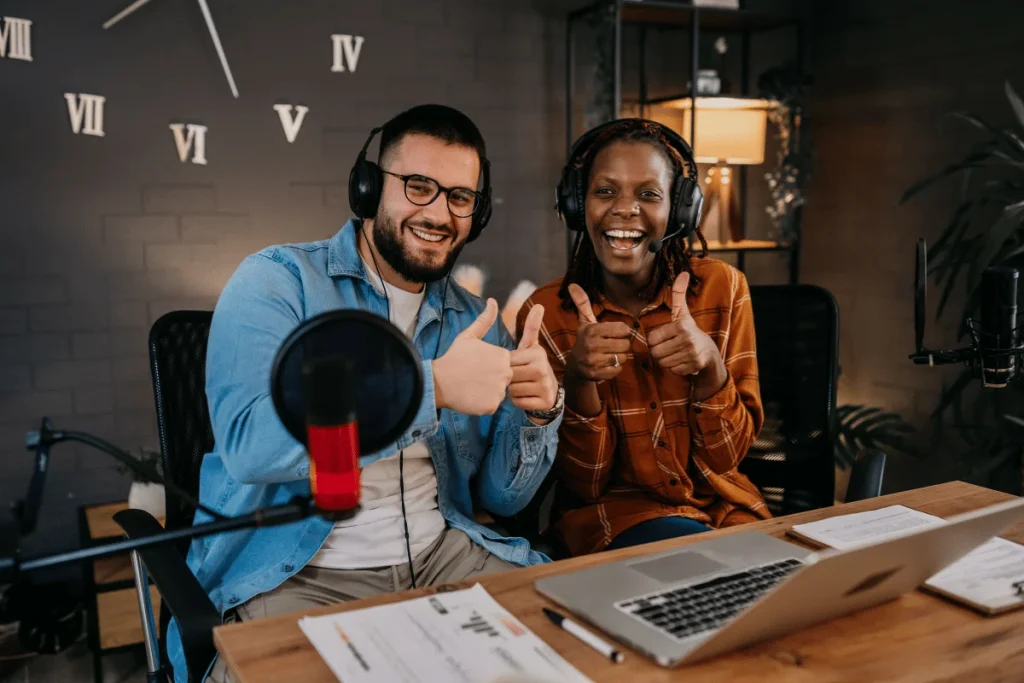 Two people in a podcast studio giving thumbs up, with microphones and laptops on the desk.