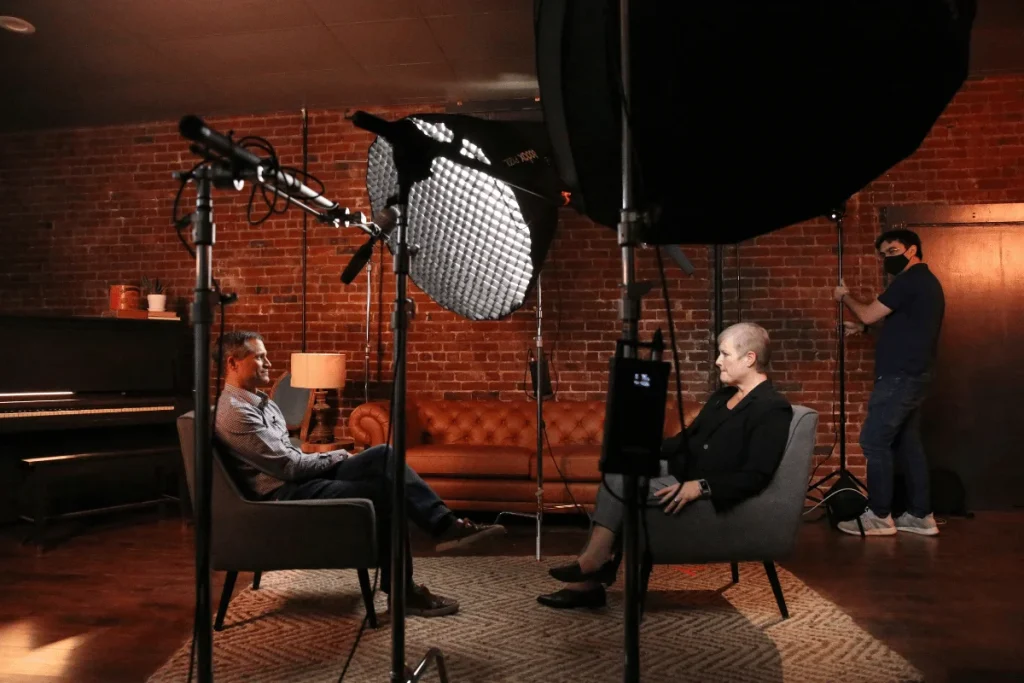 Interview setup with studio lights and two people seated facing each other, one adjusting equipment.