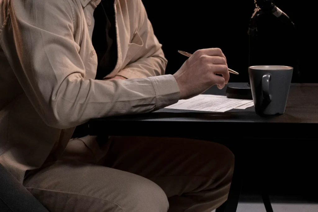 Person writing on paper at a desk with a coffee mug and microphone in dim lighting.