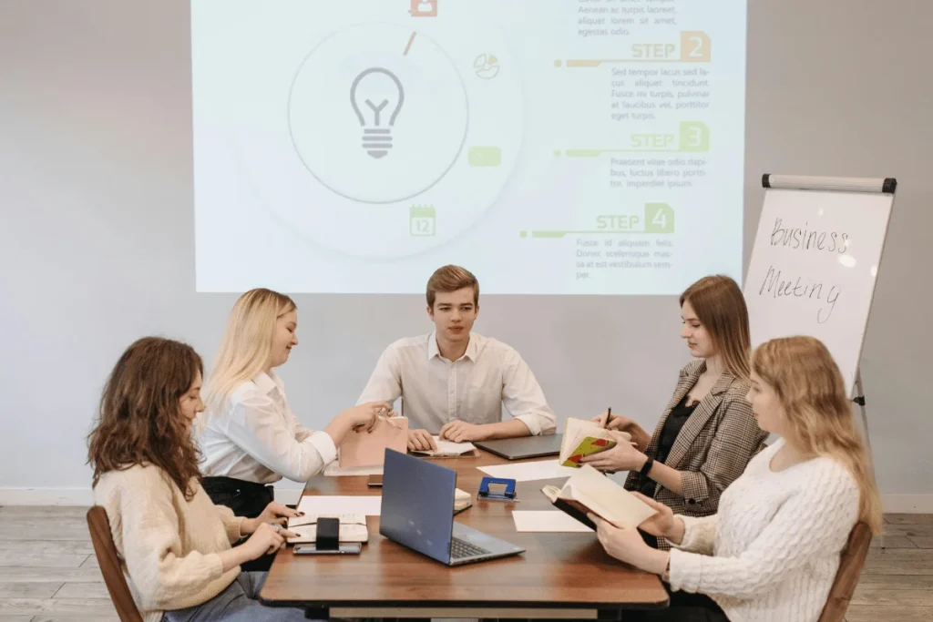 Group of professionals engaged in a meeting with project steps displayed on screen and flipchart text "business meeting."