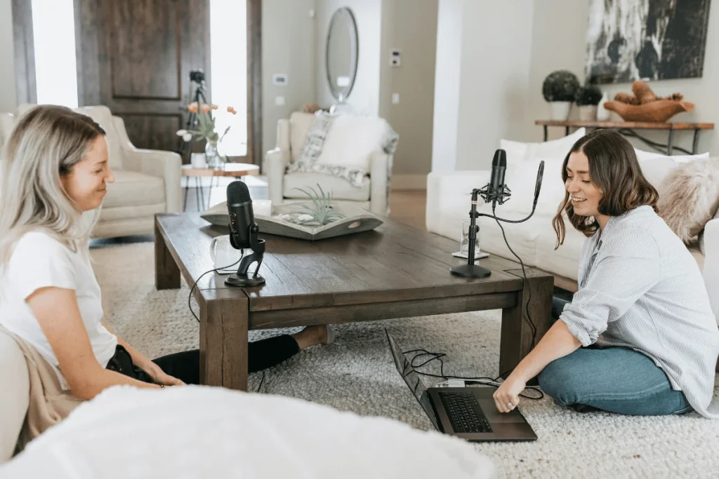 Two women recording a podcast in a cozy living room setup with microphones and a laptop.