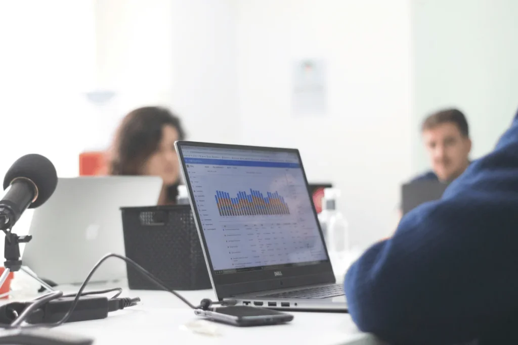 Laptop displaying bar graph analytics during a meeting with microphones and participants in the background.