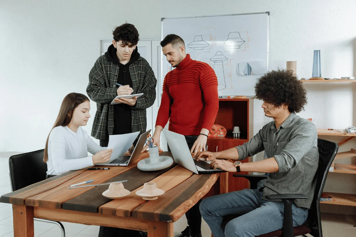 Group of four people in a meeting with laptops, papers, and design sketches on a whiteboard.