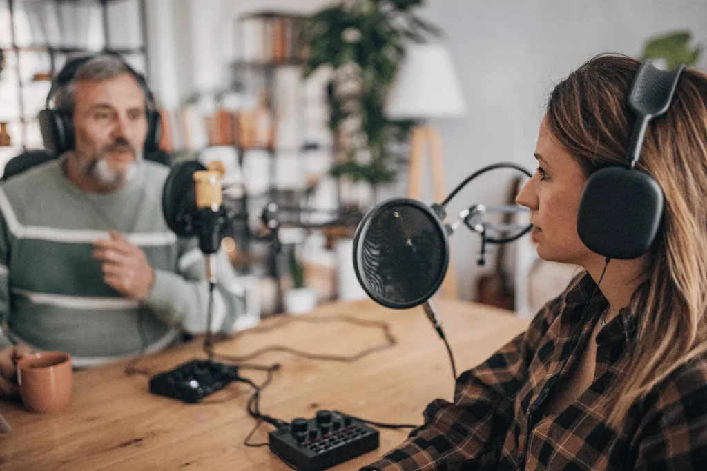 Two people with headphones speaking into microphones in a podcast recording setup.