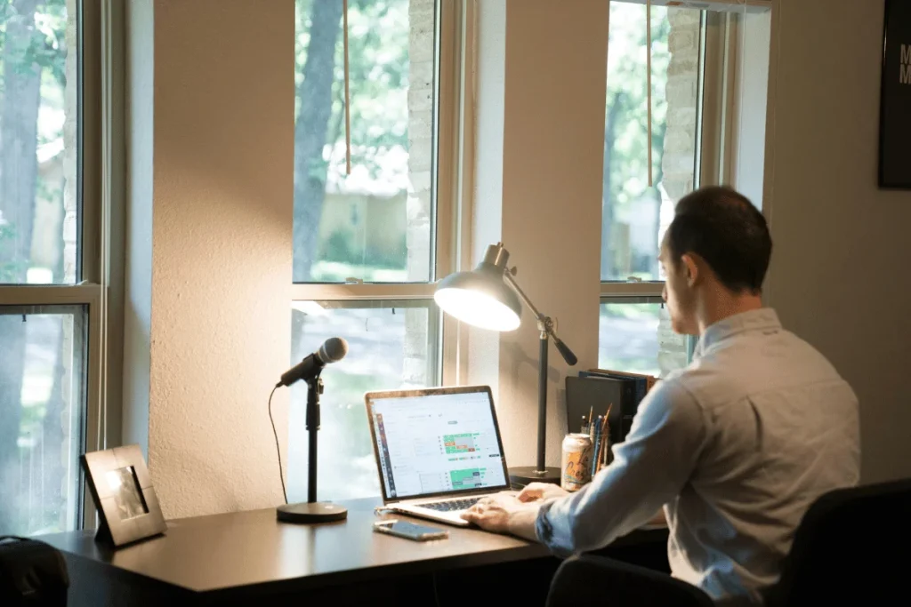 Man working on laptop at home office desk near window with desk lamp and microphone.