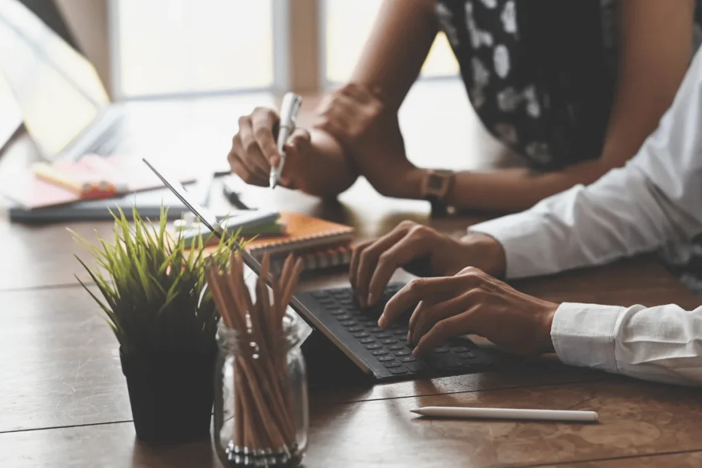 Close-up of two people working at a desk with a computer and notepad.