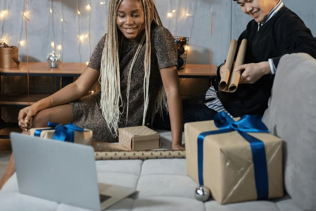 Two people wrapping gifts with ribbon and paper on a couch, festive lights in background.