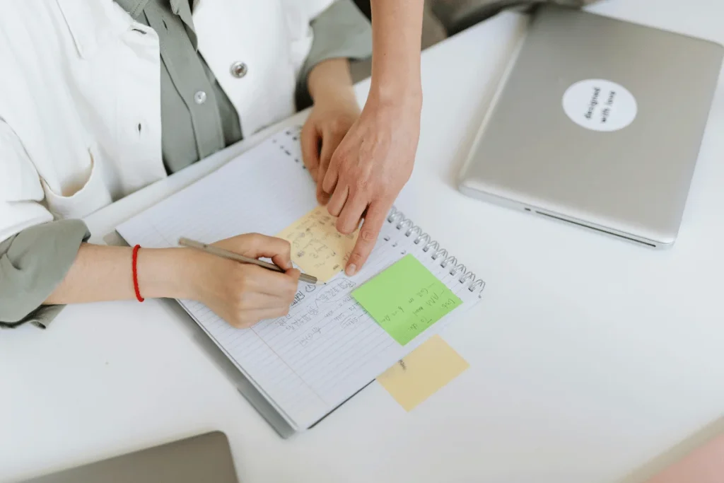 Person writing notes in a notebook with sticky notes and a laptop nearby.