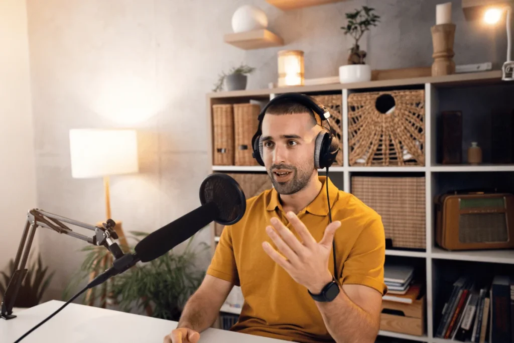 Person with headphones speaking into a microphone in a cozy room with shelves.