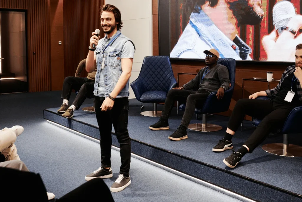 A person standing and speaking into a microphone with seated attendees in a conference room.