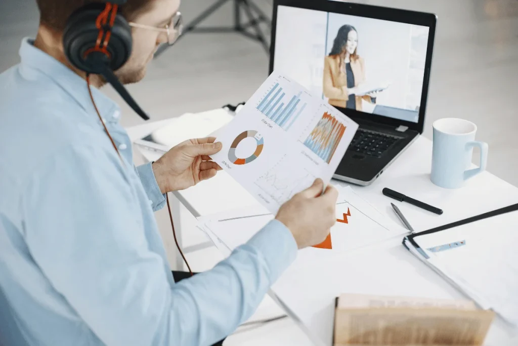 Person analyzing graphs during a video call with a laptop and coffee cup on desk.