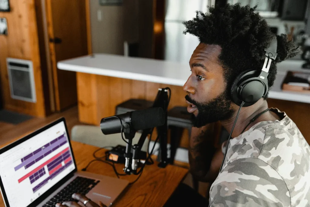 Person with headphones in front of a microphone and laptop, suggesting podcast recording or streaming.