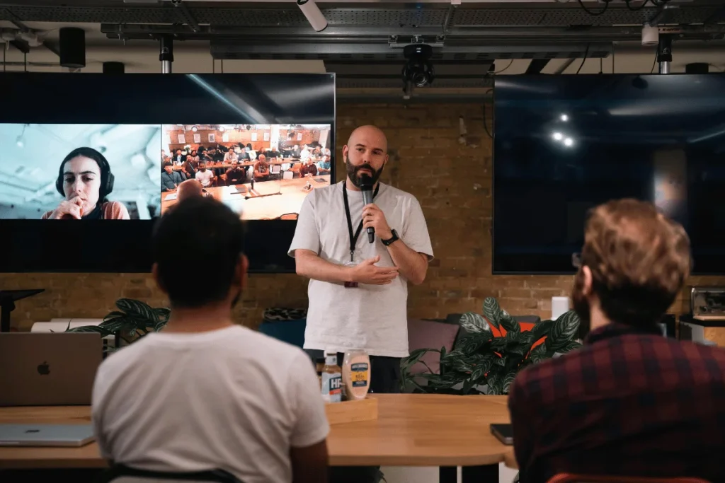 Man giving a speech at an event with a video call displayed on screens.