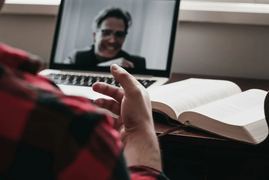 A person attending a virtual meeting on a laptop with a book open on the desk.