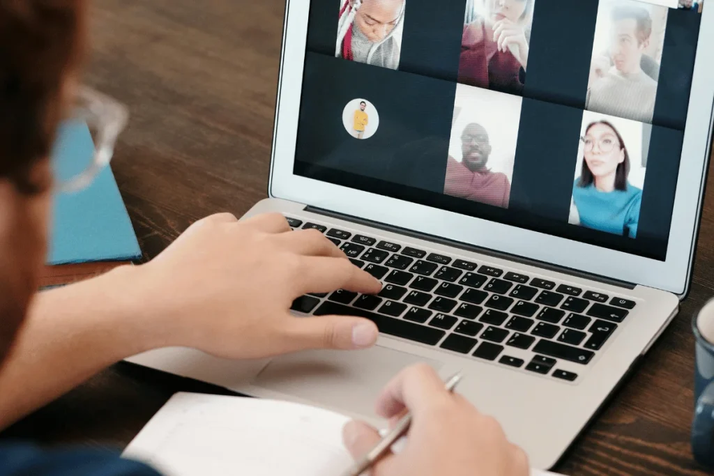 Person on a video call with multiple participants on a laptop, taking notes.