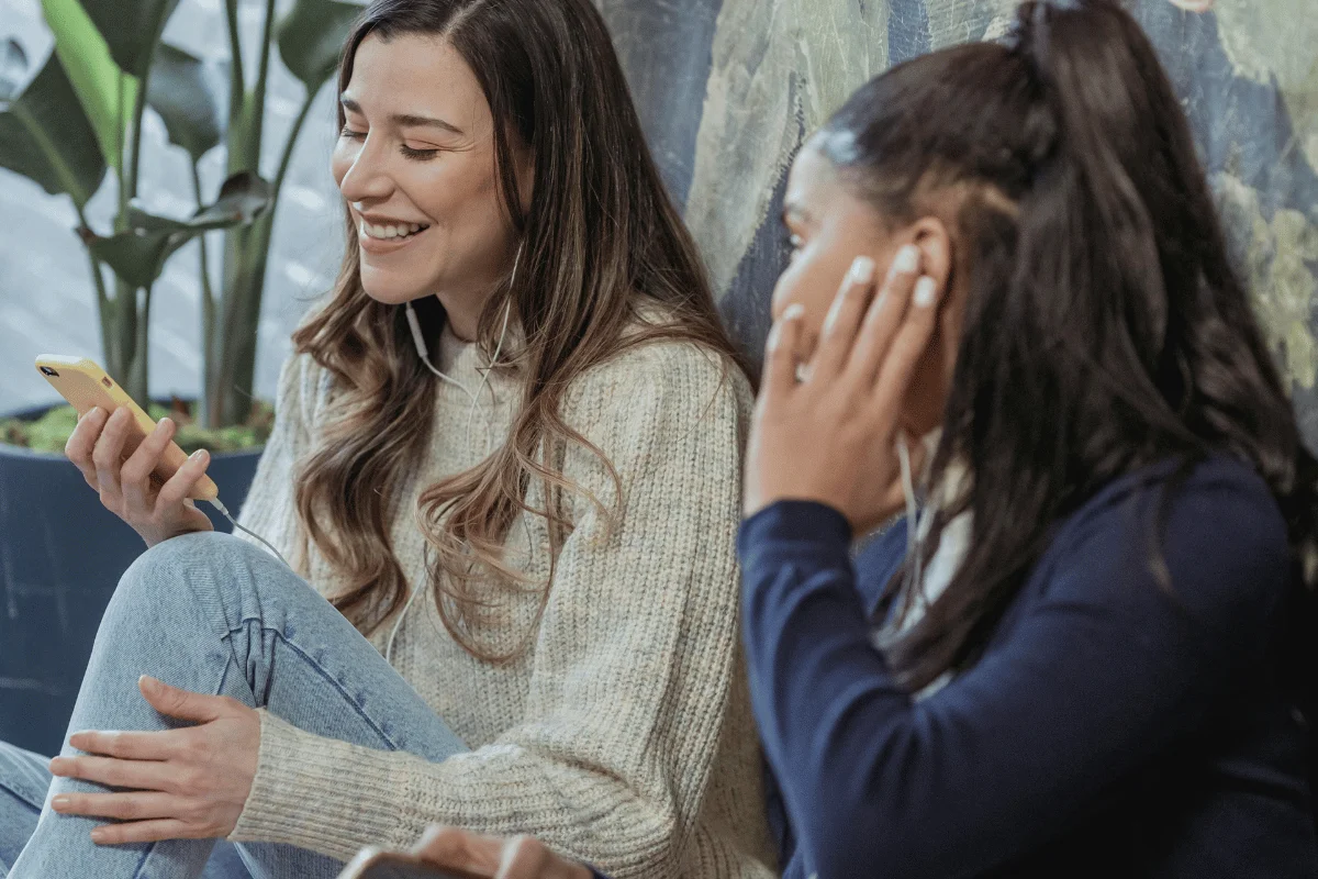 Smiling woman enjoying content on phone beside friend with earphones in cozy indoor setting.