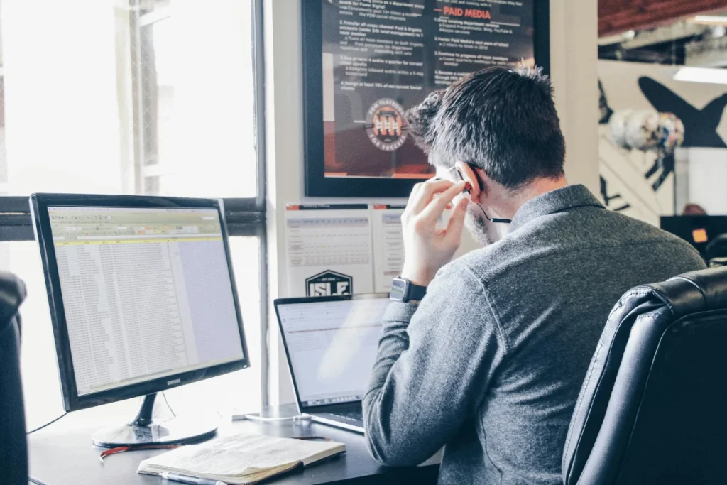 A man in a grey sweater sits at a desk, focused on two computer screens while wearing a headset and holding his glasses.