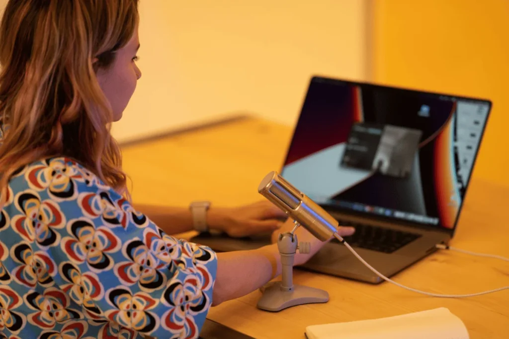 Podcast host using a desktop microphone and laptop in a brightly lit workspace.