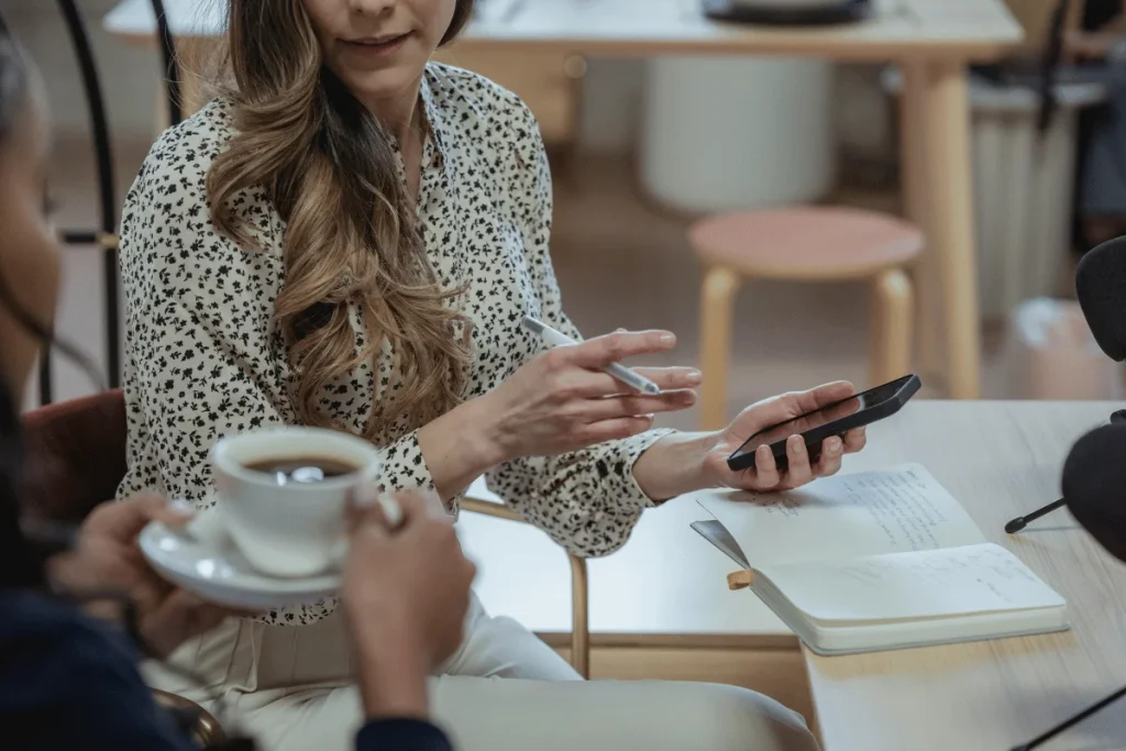 Woman holding a smartphone and discussing ideas while her colleague enjoys coffee during a casual meeting.