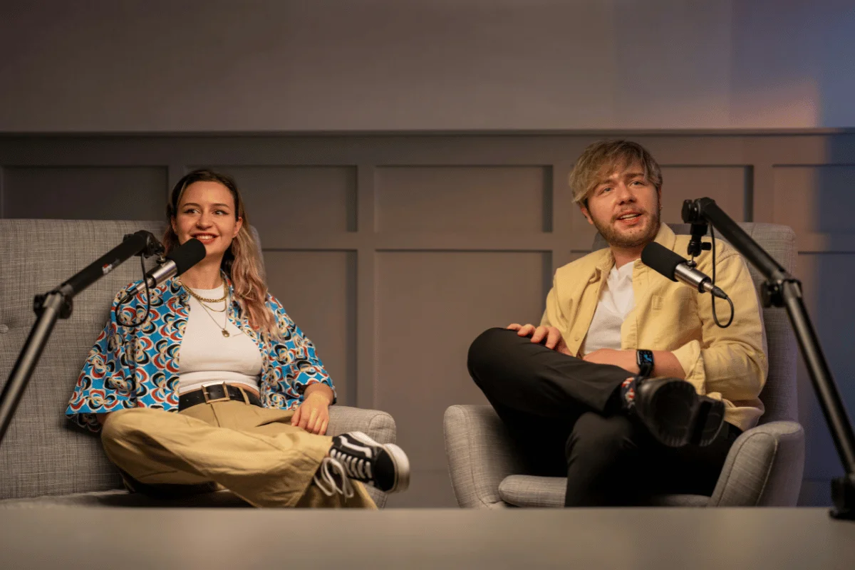 Two podcast hosts sitting in armchairs, speaking into professional microphones.