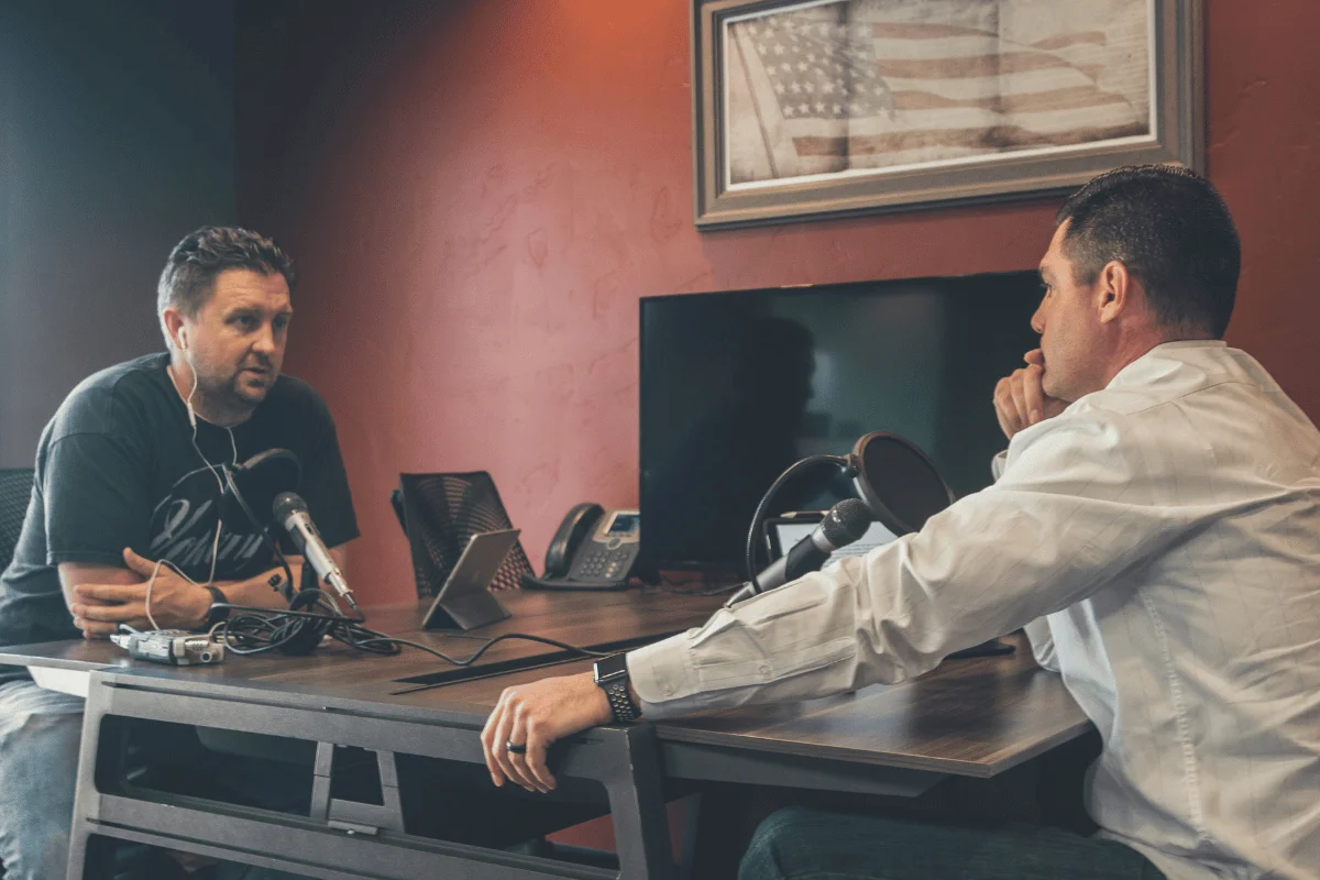 Two men engaged in a podcast recording session with microphones on a wooden table in a modern studio.