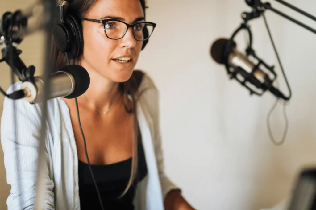 Woman wearing glasses and headphones, speaking into a microphone during a podcast recording.