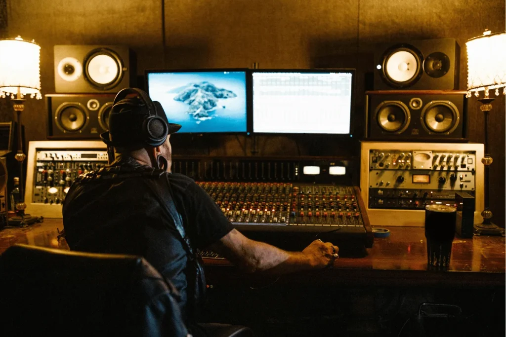 Music producer in recording studio adjusting audio levels on a mixing console, surrounded by speakers and monitors.