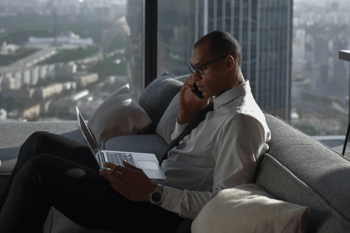 A man in a white shirt and tie sits on a sofa, using a laptop with a city skyline visible through large windows.