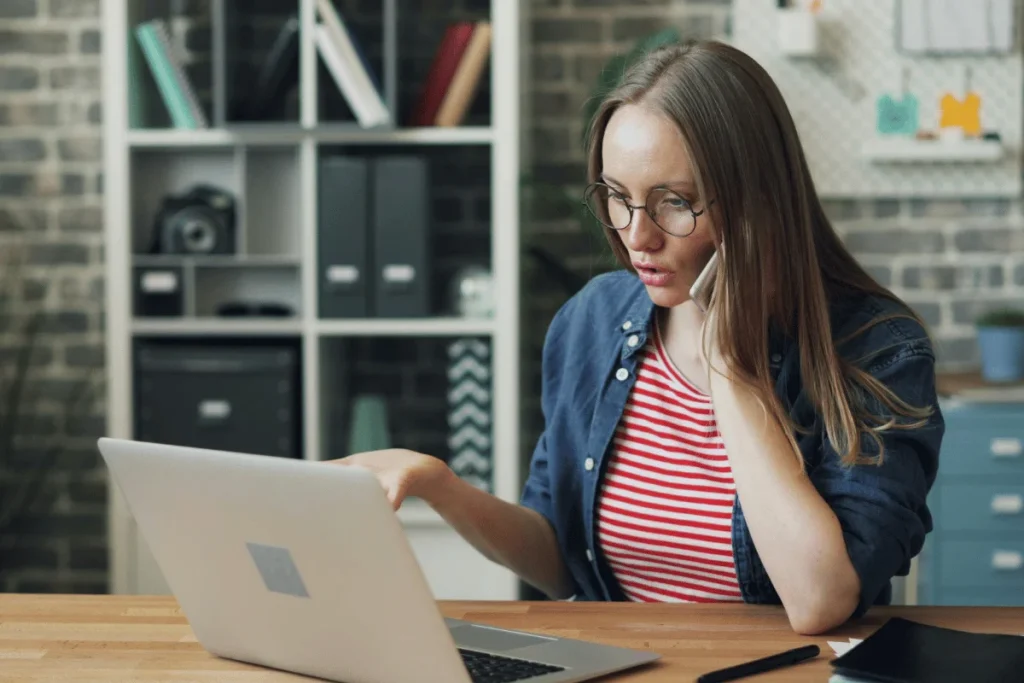 Young woman working remotely on a laptop while talking on the phone.