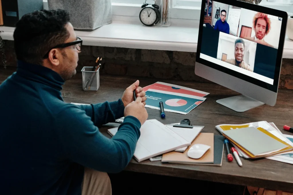 A person is seated at a desk, focused on a video conference call displayed on a large monitor, surrounded by office supplies.