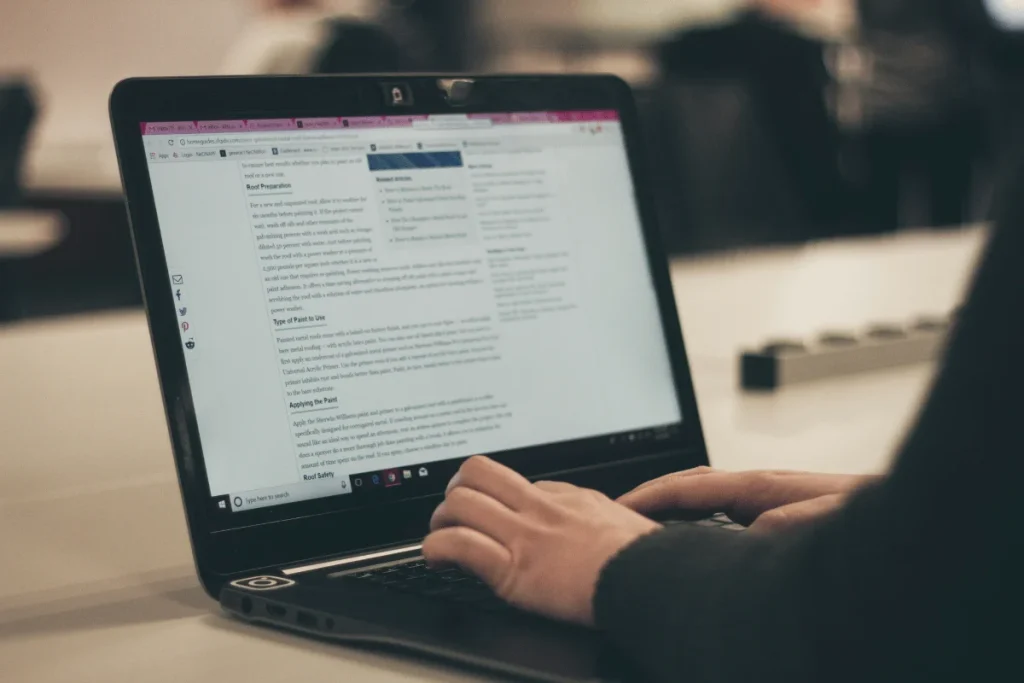 A person typing on a laptop displaying an article about roof preparation and painting techniques in a well-lit workspace.