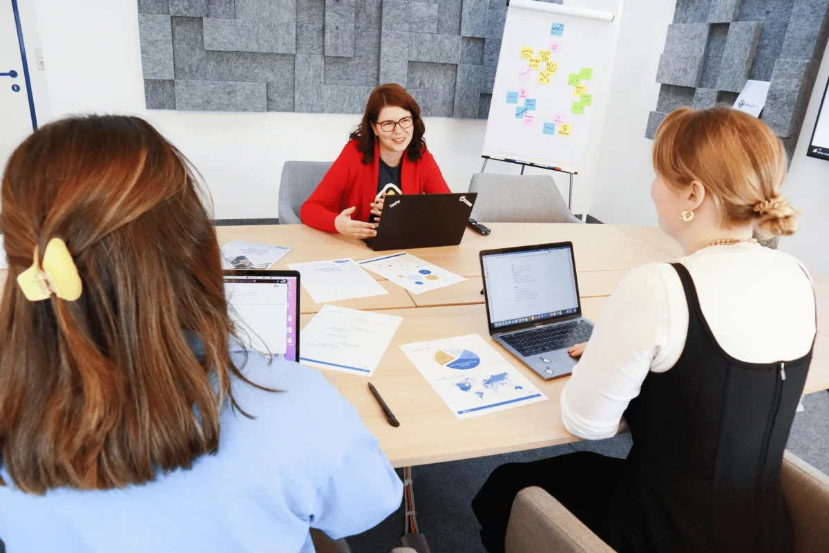 Three women engage in a collaborative meeting around a table with laptops, notes, and a whiteboard filled with colorful sticky notes.