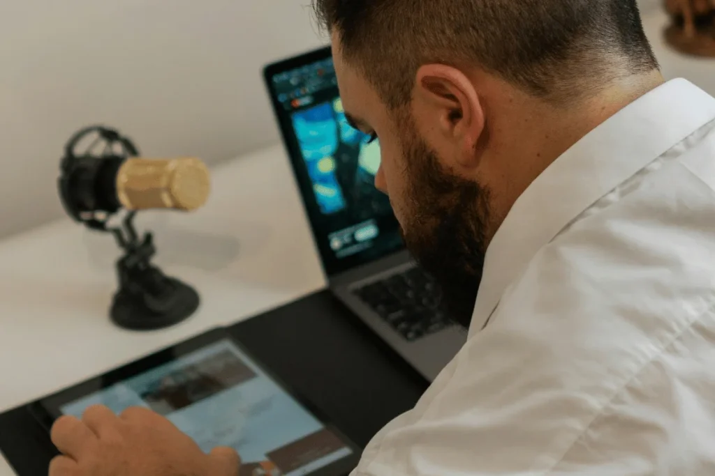 A man working with a tablet and laptop, accompanied by a gold microphone on a desk setup.