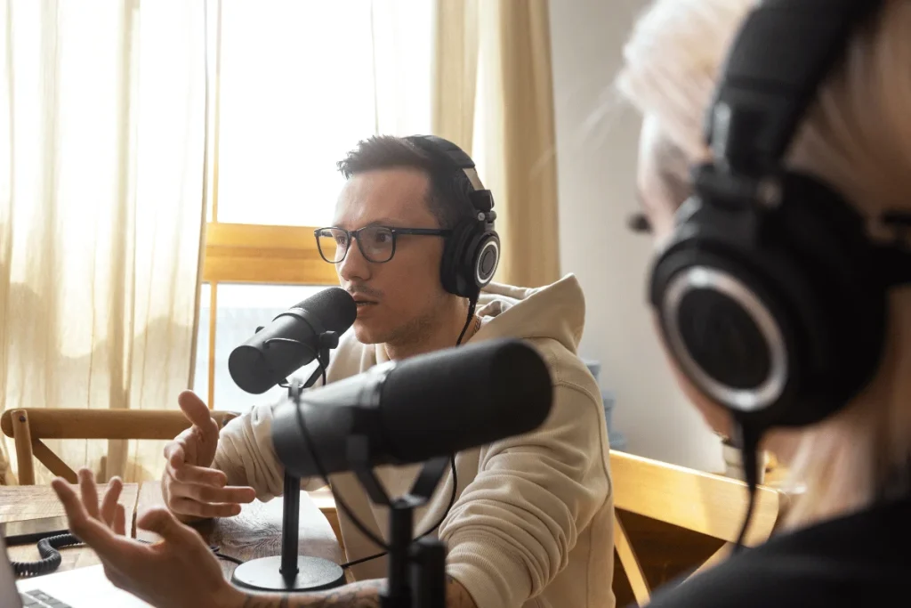 Two people are engaged in a podcast discussion, wearing headphones and speaking into microphones, with soft natural light in the background.