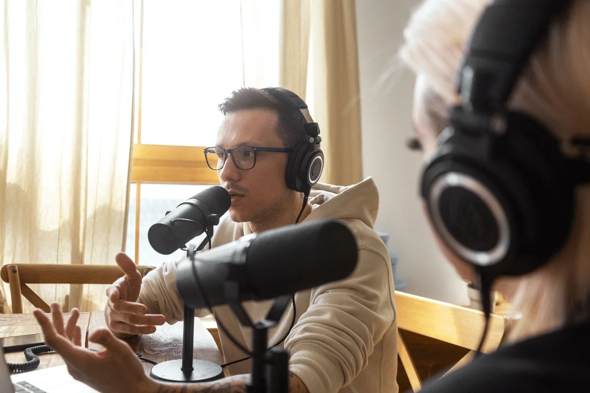 Two people are engaged in a podcast discussion, wearing headphones and speaking into microphones, with soft natural light in the background.