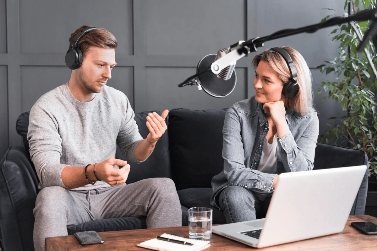 A man and a woman in headphones are engaged in conversation on a couch, with a laptop and a microphone in front of them.