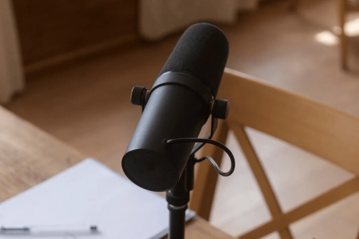 A close-up of a black microphone positioned on a stand, with a wooden table and paper visible in the background.