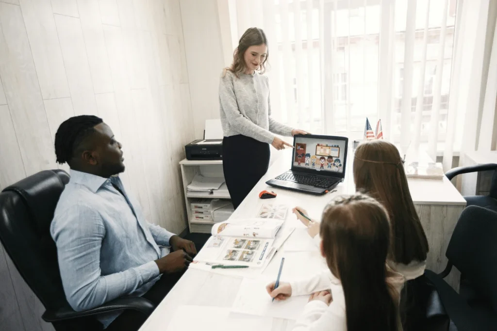 A woman pointing at a laptop screen while explaining something to a group of people in an office setting.